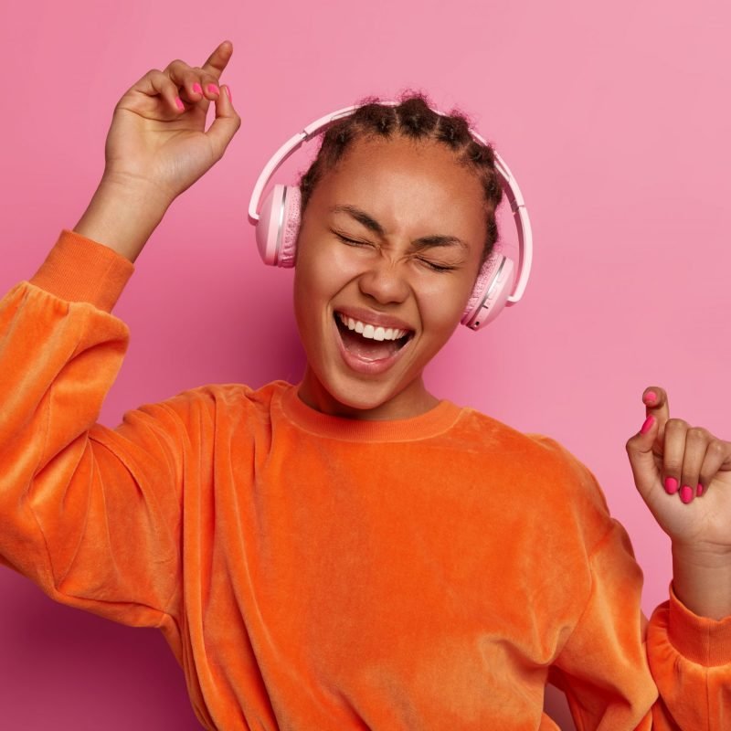Dance with me. Upbeat dark skinned teenager raises arms, moves with rthythm of melody, feels energetic, dressed in bright orange jumper, has fun, isolated on pink background. Music brings joy
