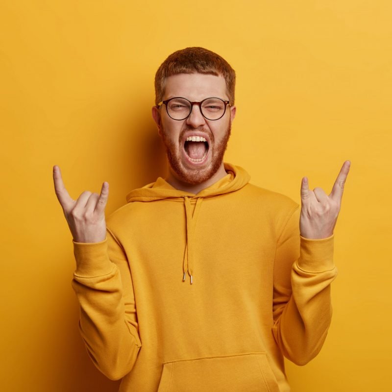Emotional teenager excliams with joy, makes rock n roll gesture, brings positive vibes, enjoys cool concert, listens favourite music, dressed in hoodie, isolated over yellow background. Body language