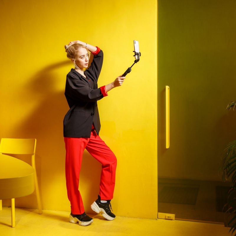 Fashion young girl blogger dressed in red trousers and black jacket takes a selfie on her smartphone in the room with yellow walls and furniture .